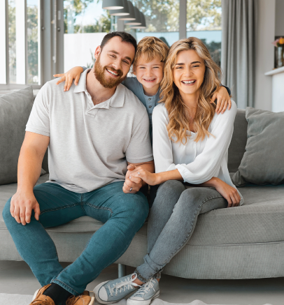 Family of three smiles on a gray sofa in a brightly lit room.