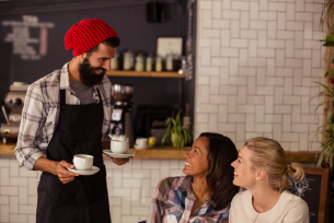 A barista in a red hat serves coffee to two smiling customers in a cafe.