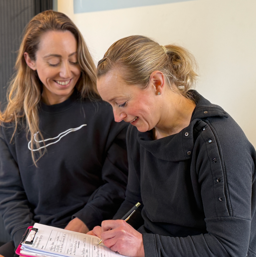 Two women are sitting next to each other writing on a clipboard