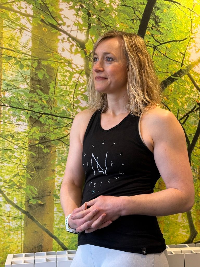 A woman in a black tank top is standing in front of a wall with trees on it.