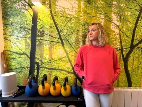 A woman in a red sweatshirt is standing in front of a wall with trees on it.
