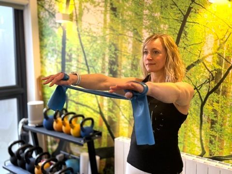 A woman is holding a blue resistance band over her shoulder in a gym.
