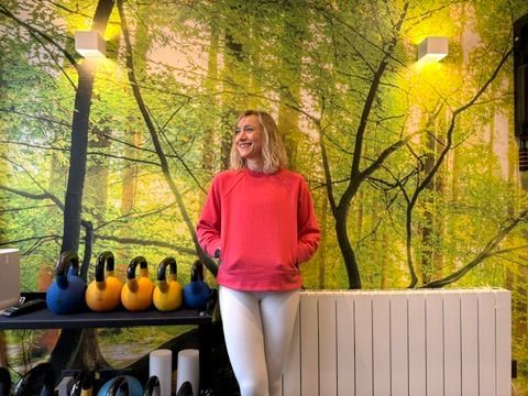 A woman in a pink sweatshirt is standing in front of a wall with trees on it.