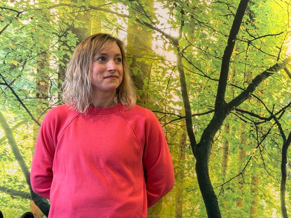 A woman in a pink sweater is standing in front of a wall with trees on it.