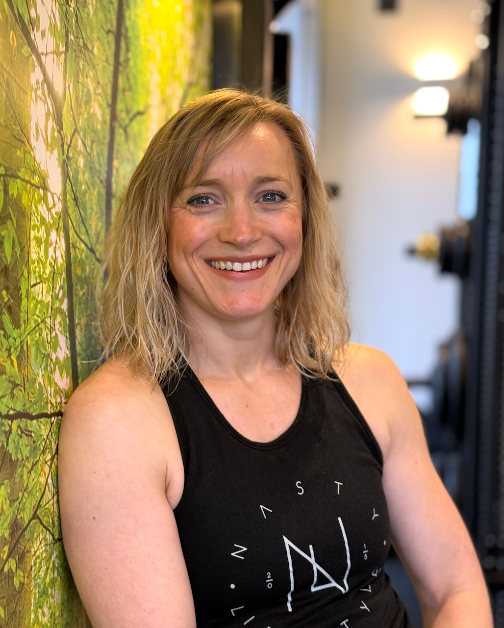 A woman in a black tank top is smiling and leaning against a wall.