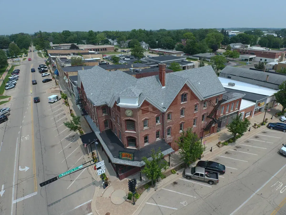 Aerial view of a brick building with a gray roof, located on a town street lined with businesses and cars.