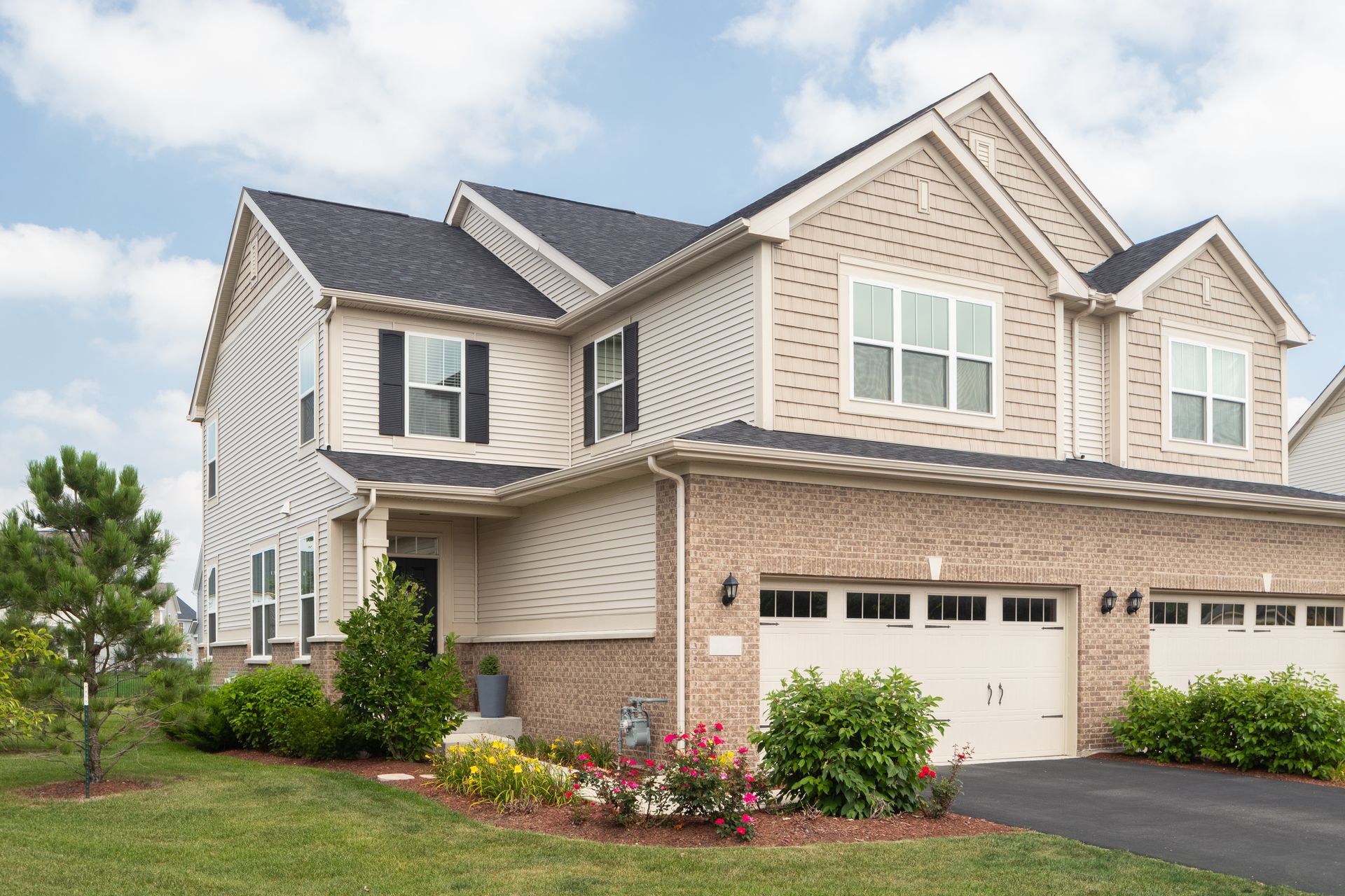 Two-story house with beige siding and brick, black shutters, garage, and green lawn under a blue sky.