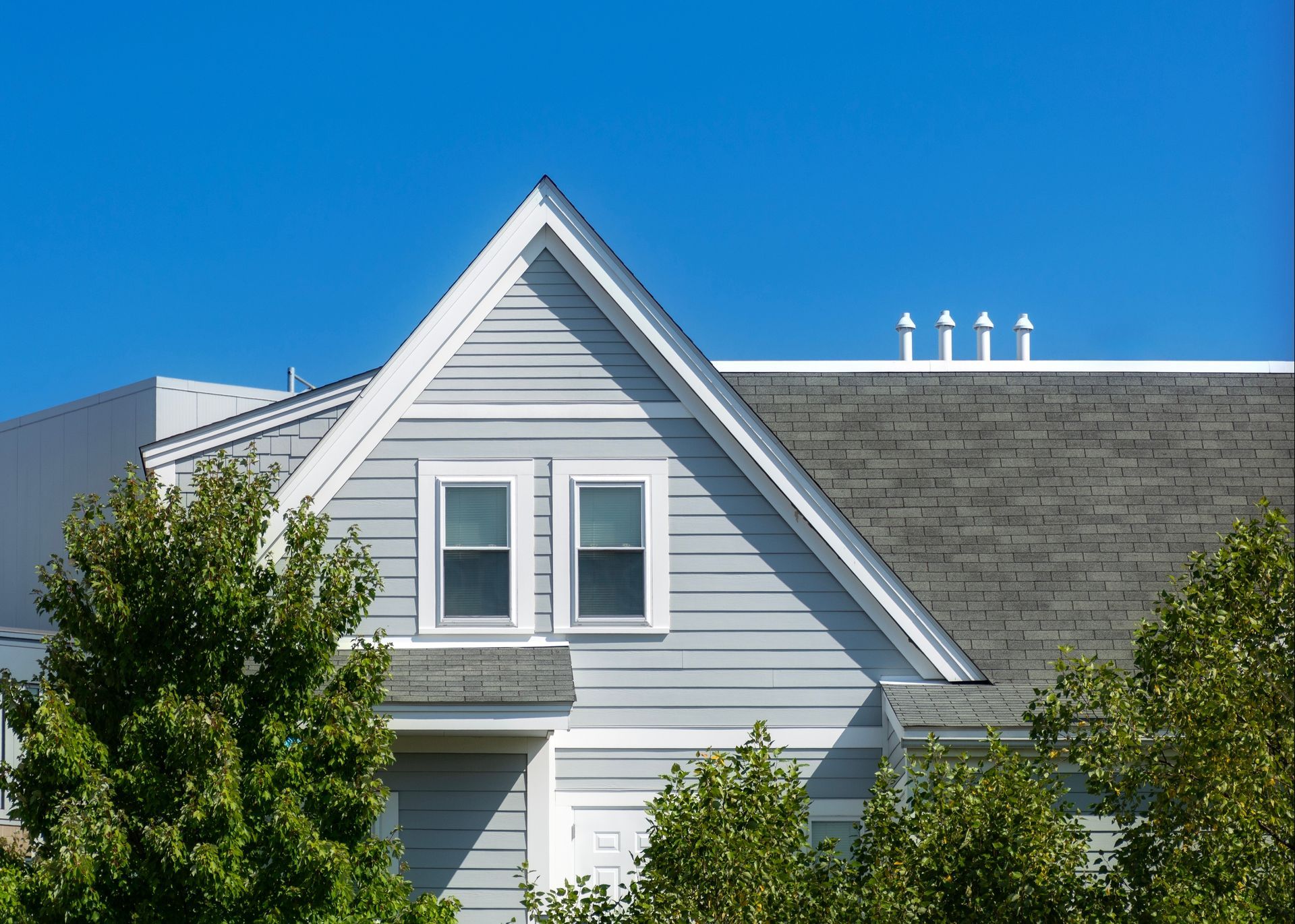 Light blue house with two windows, gray roof, and green trees against a blue sky.