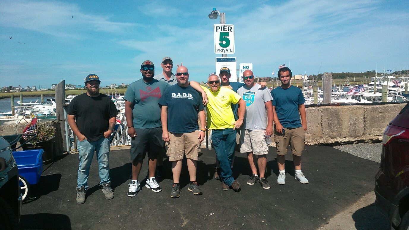 Group of men posing near boats, some smiling or gesturing. Outdoors, cloudy sky.