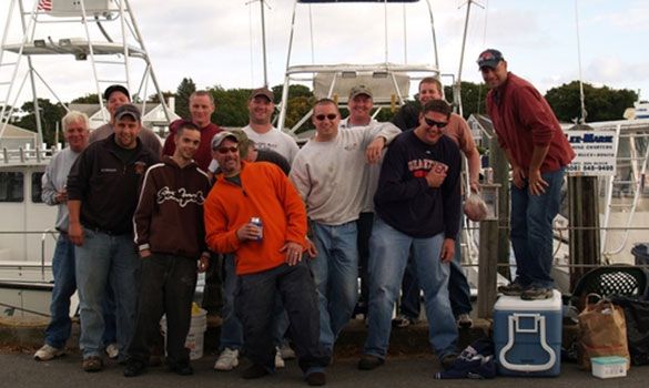 Group of men posing near boats, some smiling or gesturing. Outdoors, cloudy sky.