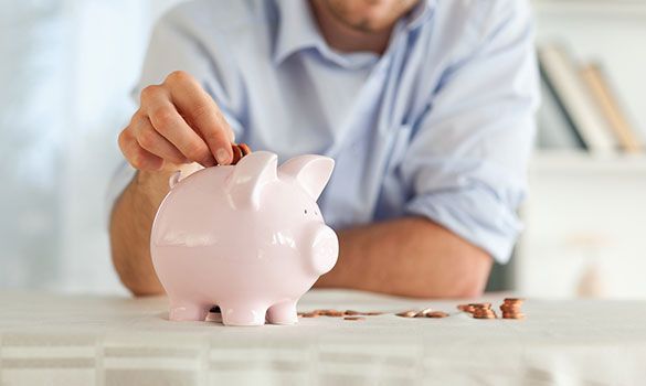 Person putting a coin into a pink piggy bank on a table with scattered coins.
