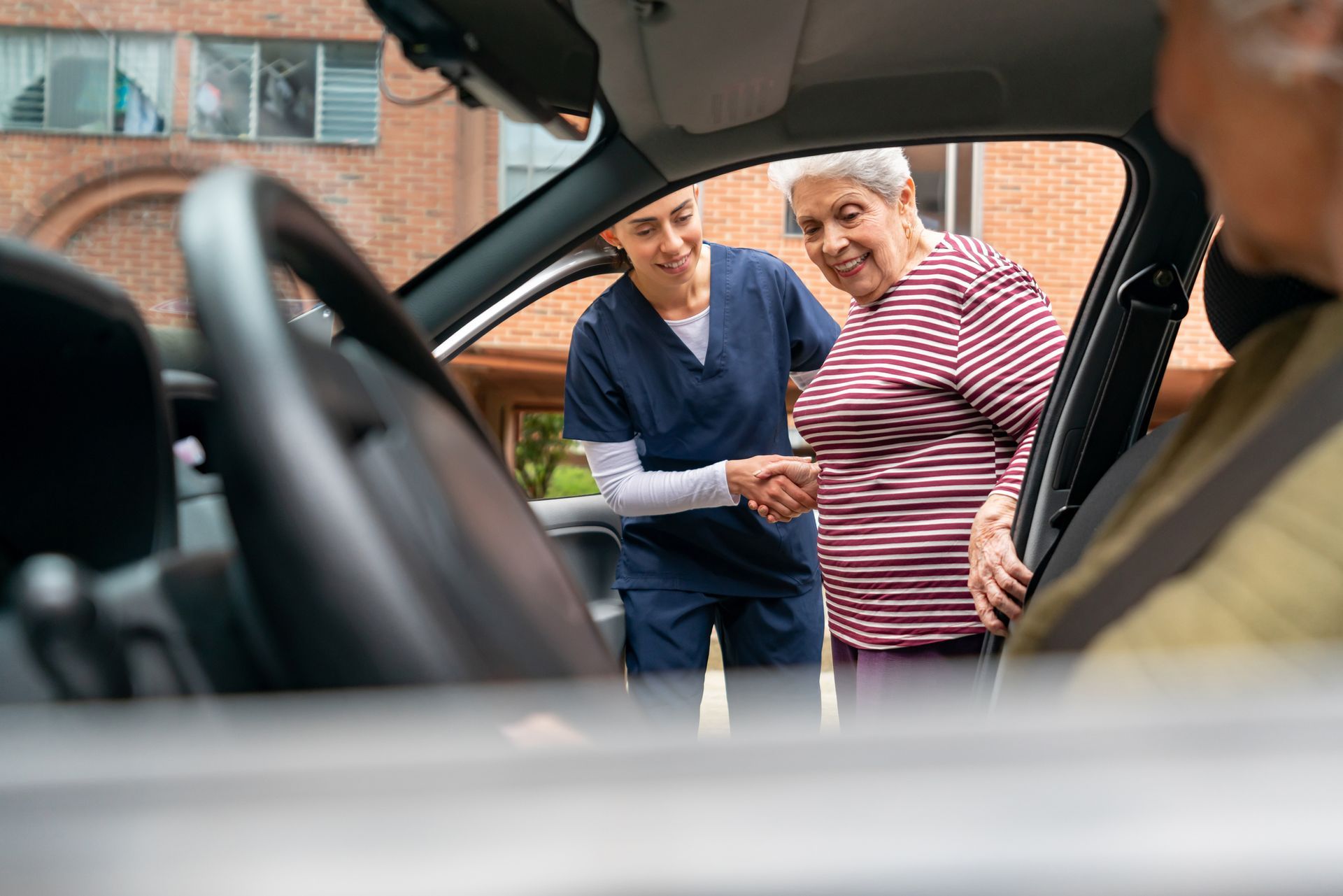 Caregiver helping a person get into a car. Both smiling. Brick building in the background.