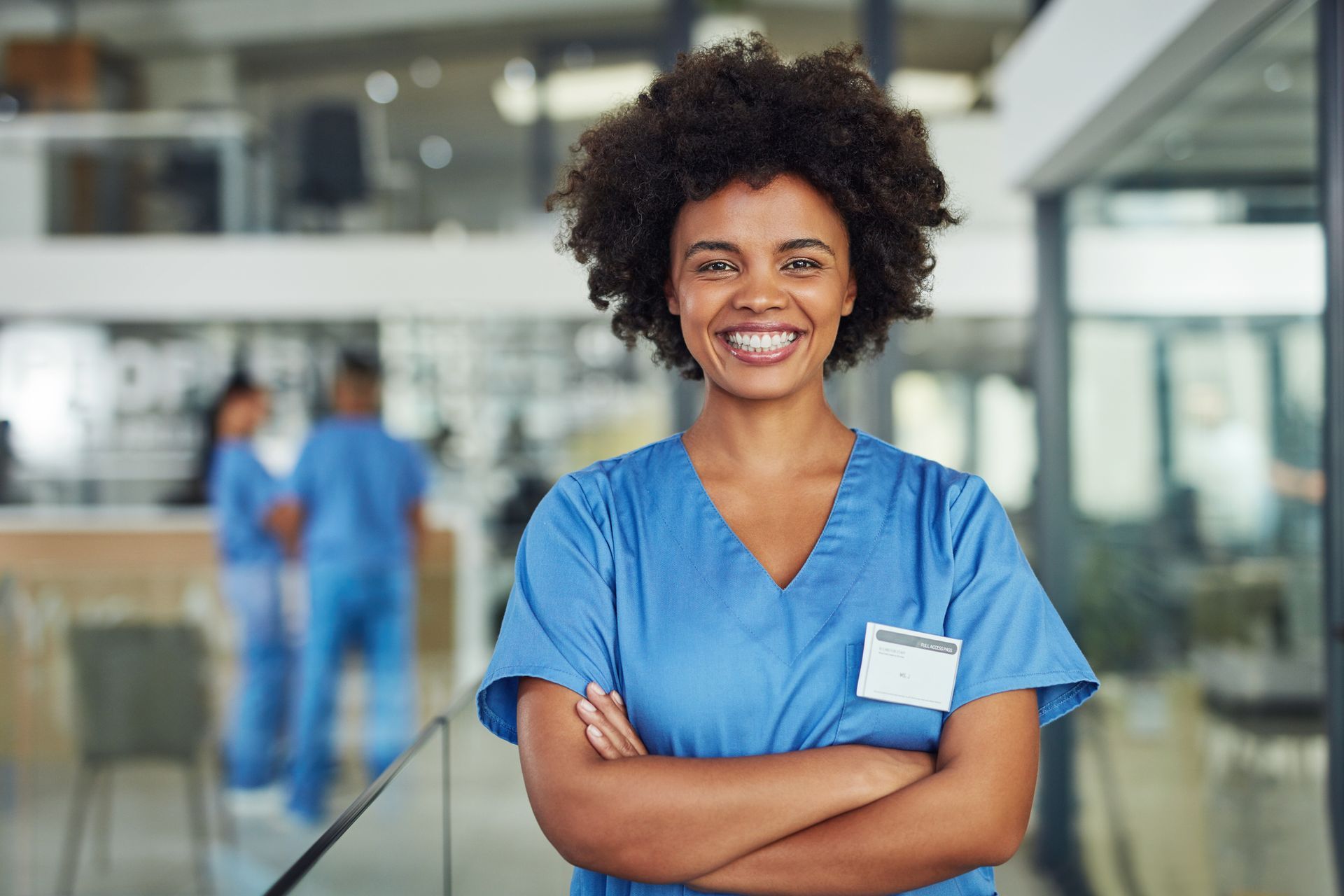 Nurse in blue scrubs with arms crossed, smiling at camera in a hospital hallway.