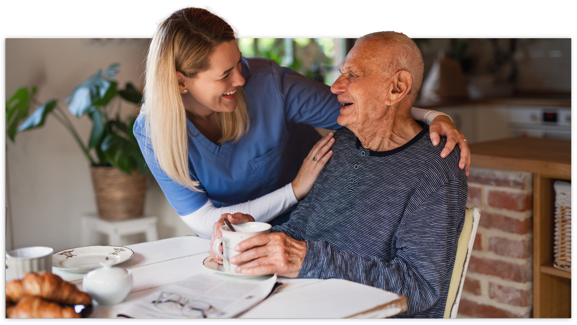 Woman in blue scrubs smiles at older person, arm around him, at a table with a mug and croissants.