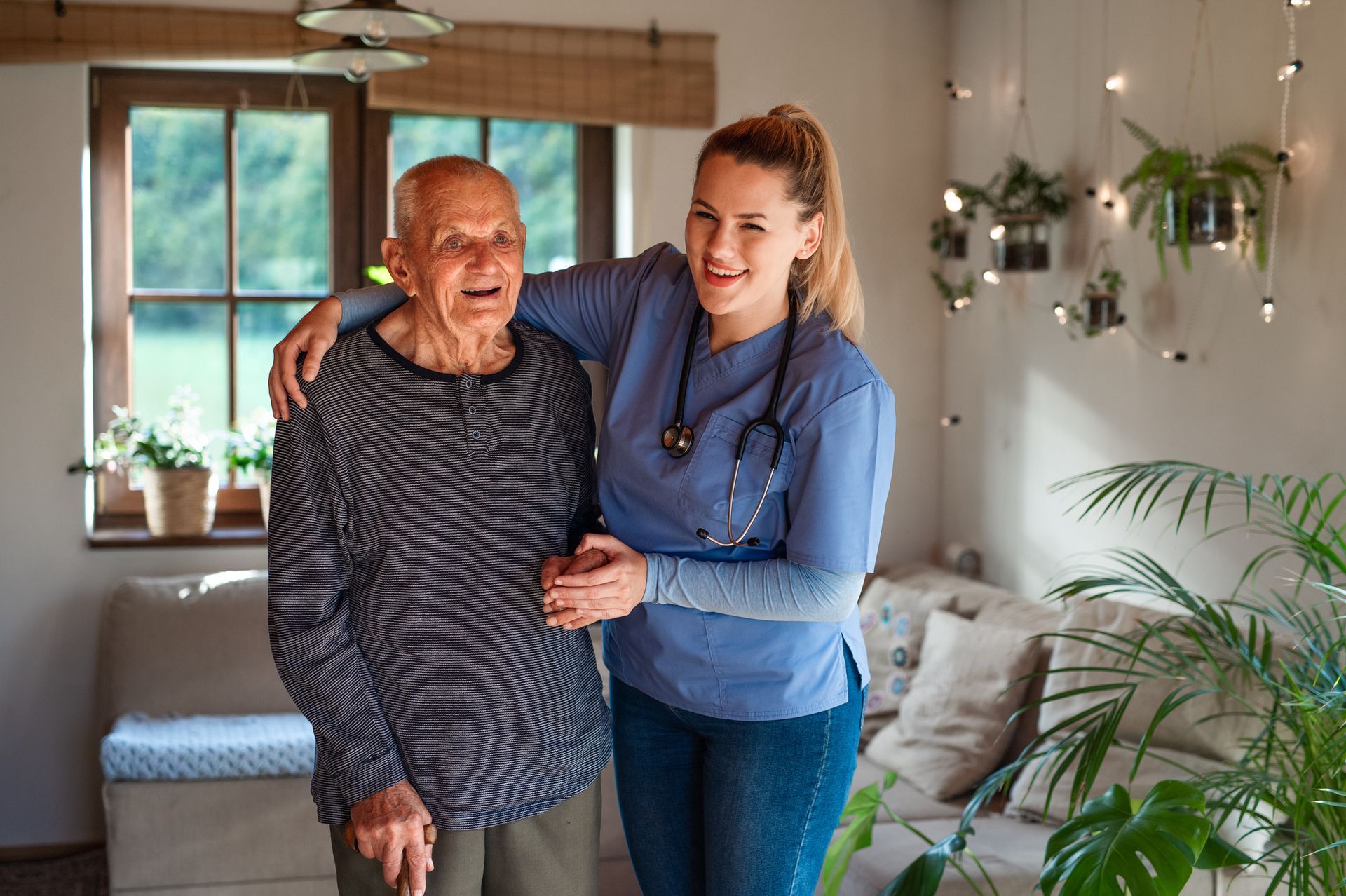 Woman in blue scrubs smiles with her arm around an older person indoors. They are both smiling.