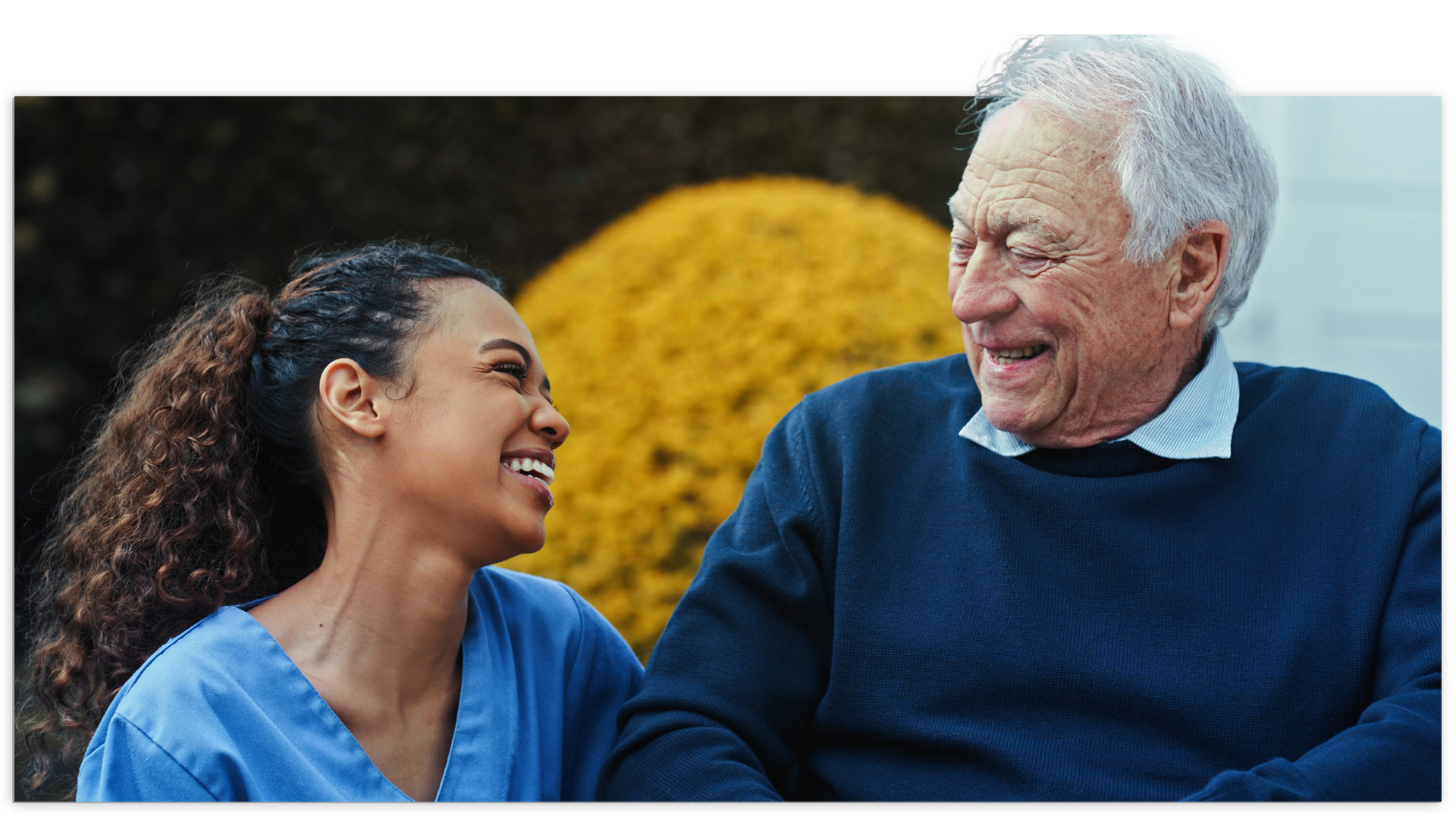 Woman in blue scrubs smiles at older man in a blue sweater. They are outdoors with yellow flowers behind them.