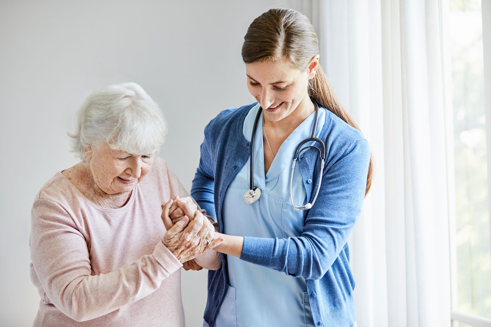 Nurse assists elderly patient with walking near a window.