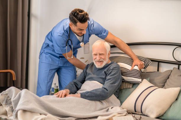 Caregiver assisting a person in bed with pillows; room setting, blue scrubs, and a smile.