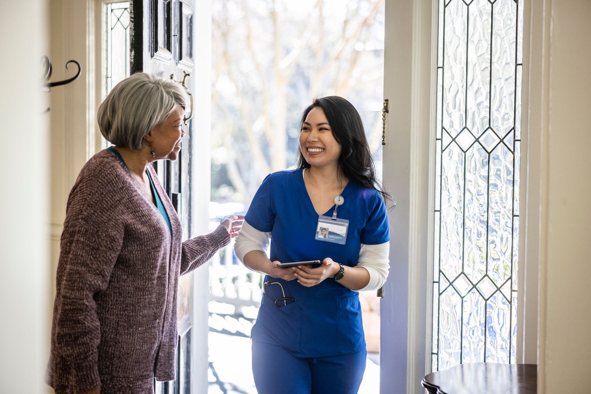 Caregiver in blue scrubs greets older woman at the doorway of a home.