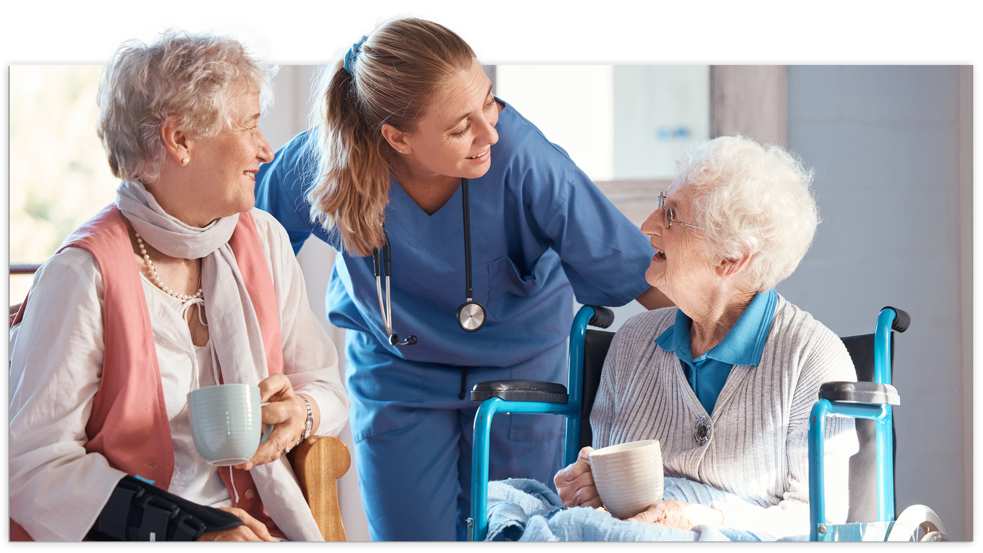 Nurse talking with two older adults, one in wheelchair, indoors; they hold mugs, smiling.