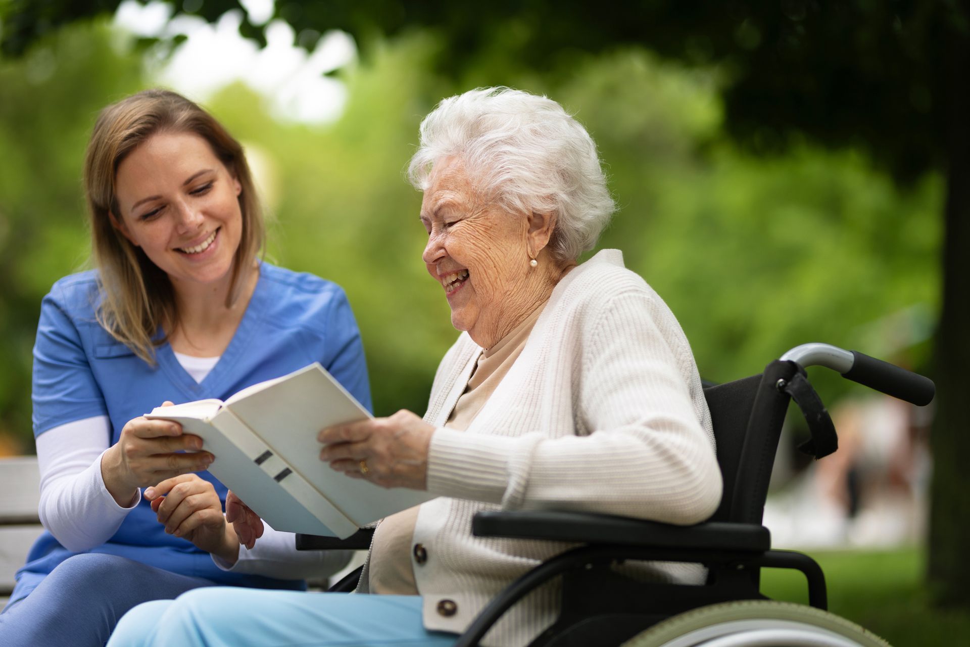 Woman in scrubs showing a book to an older person in a wheelchair; both smile outdoors.