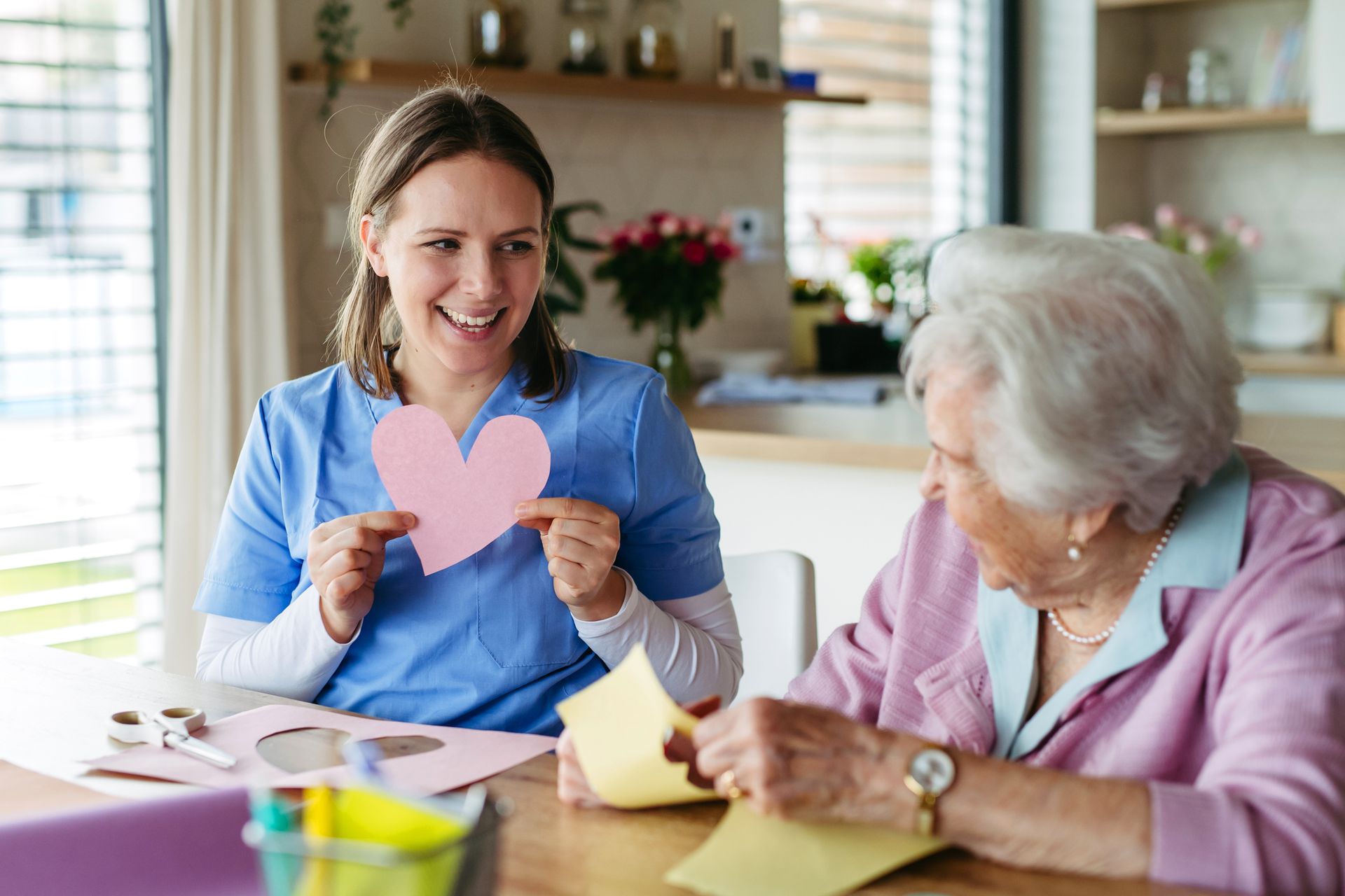 Woman in blue scrubs shows a pink heart to an elderly person crafting at a table. Bright room setting.