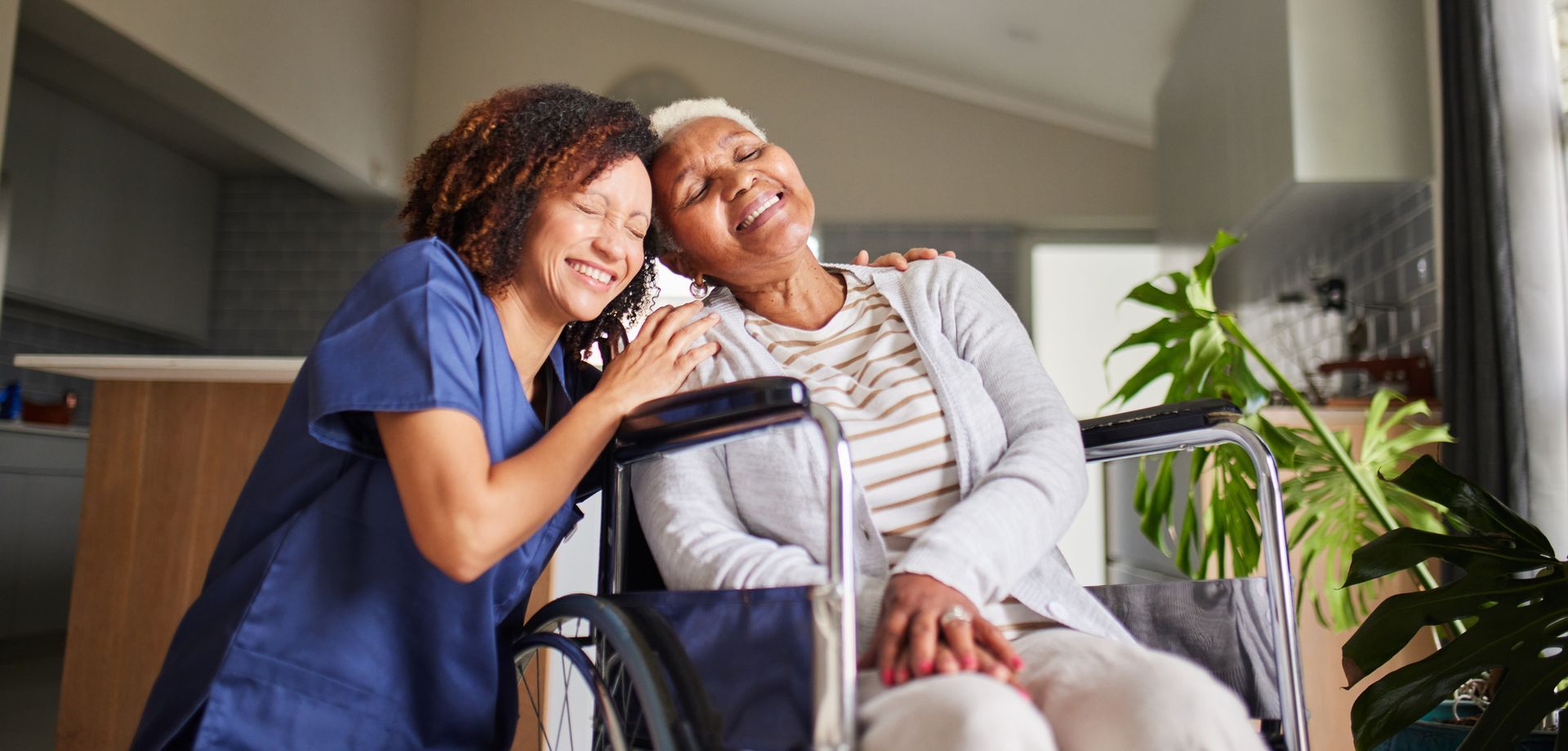 Woman in wheelchair with caregiver smiling. Inside a home setting, near a window with plants.