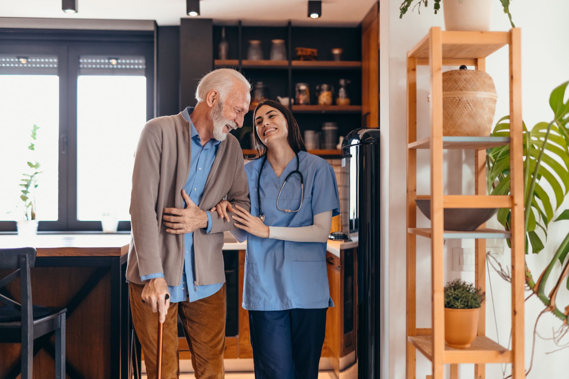 Caregiver assists senior man with a cane. They are in a well-lit home, smiling.