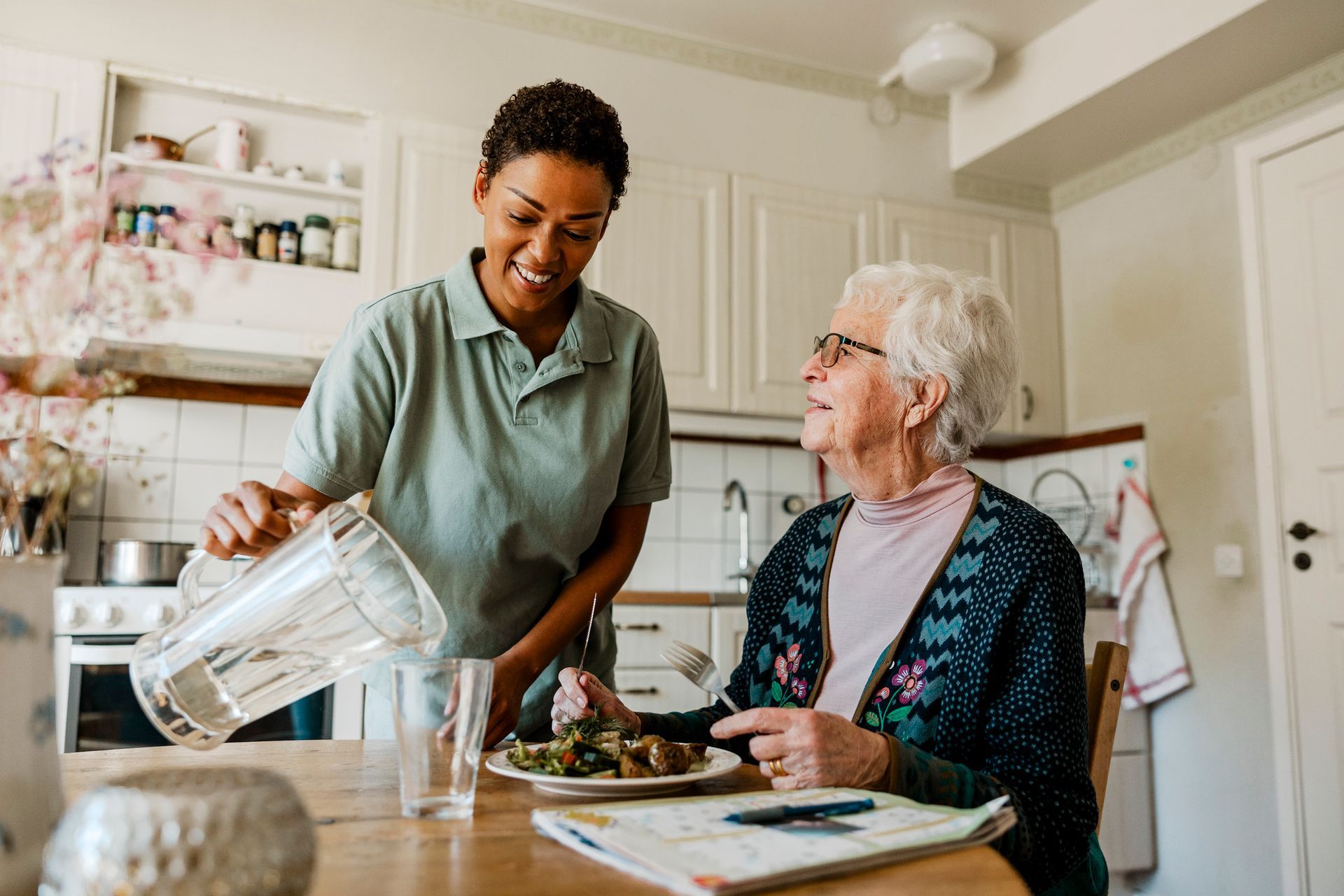 Caregiver pouring water for an elderly person at a table, both smiling. Bright kitchen setting.