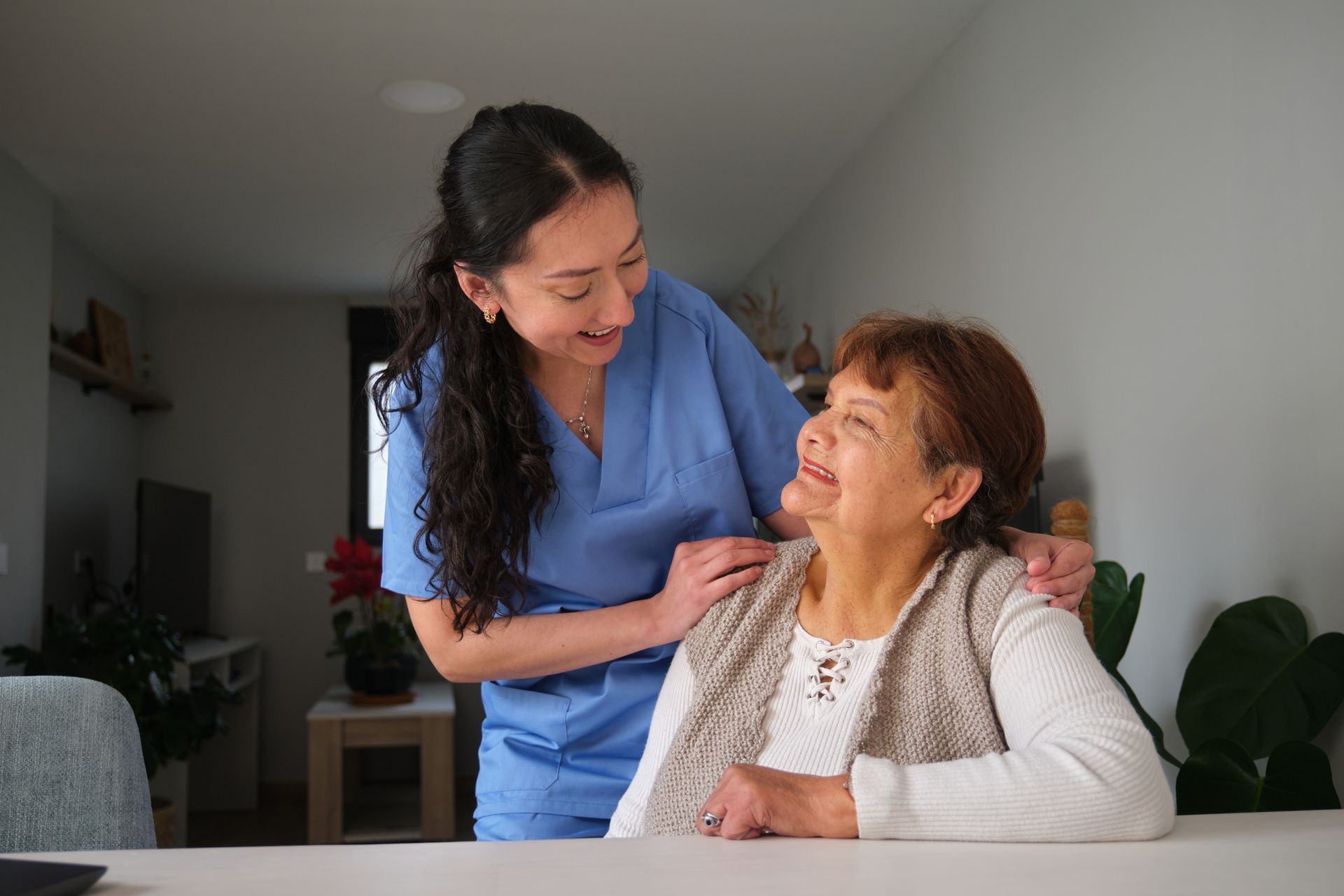Caregiver in blue scrubs smiles at elderly person, hands on her shoulders. Indoors, natural light.