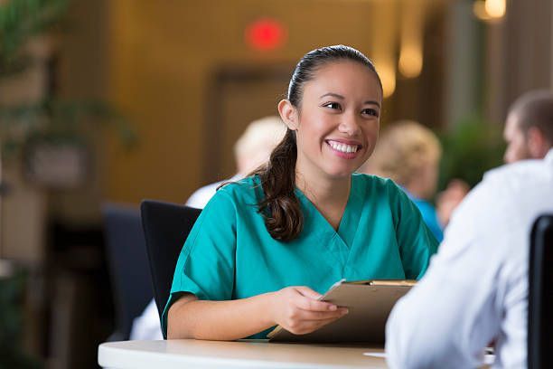 Nurse in teal scrubs smiles, holding a clipboard, talking to someone at a table indoors.