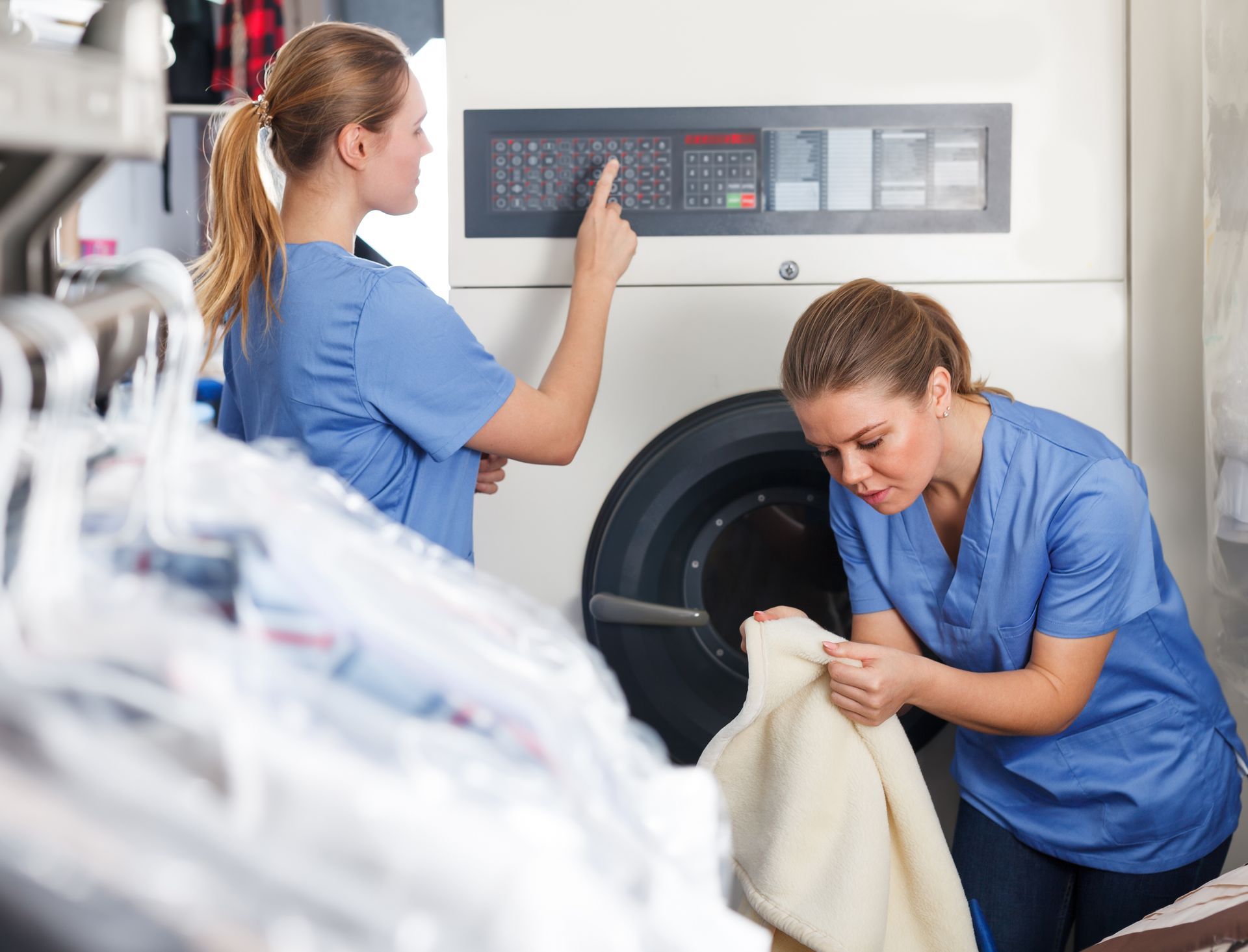 Two women in blue scrubs working in a dry cleaning facility; one operates a machine control panel, the other inspects fabric.