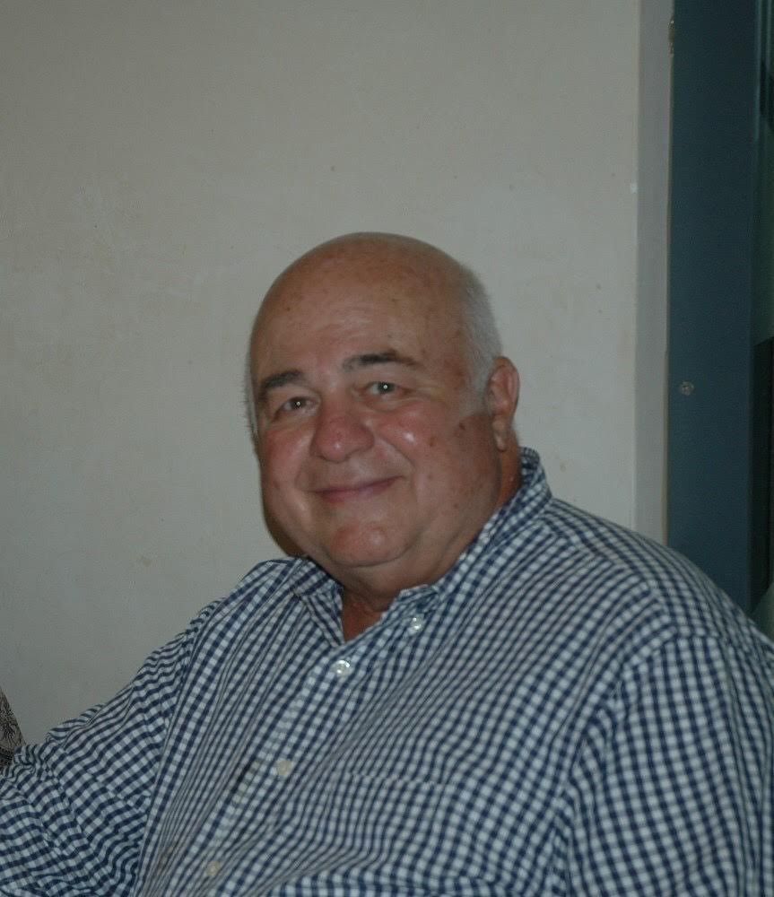 A bald man smiles, wearing a blue and white checkered shirt. He is indoors.