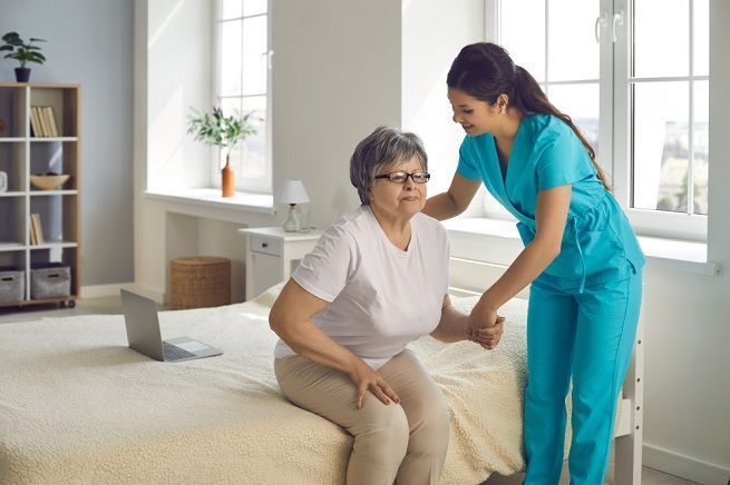 Caregiver assists a person to sit up on a bed. Bright room, window, neutral colors.