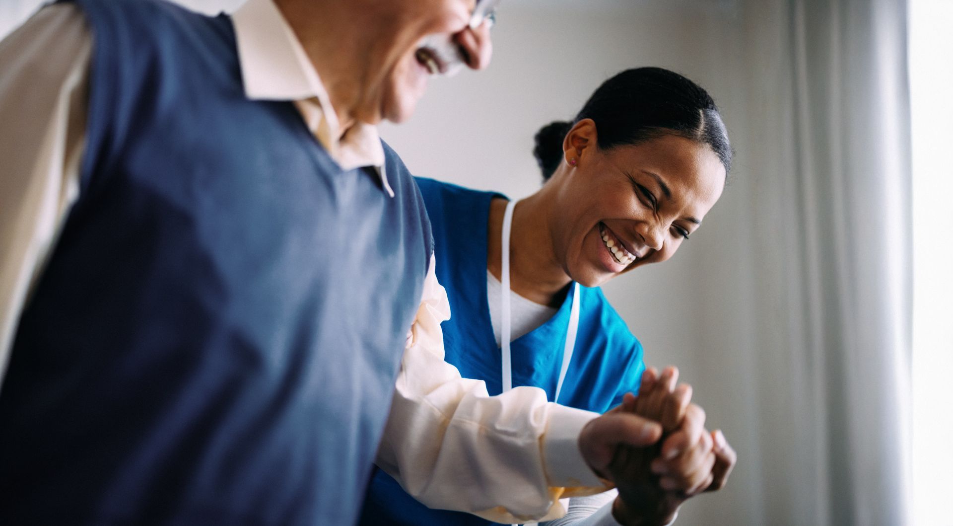 Nurse helping a person walk, both smiling. The nurse is wearing blue scrubs.