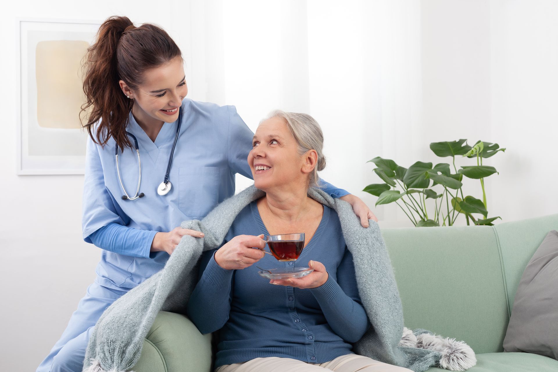 Nurse comforting an older person with a blanket and cup of tea on a couch.