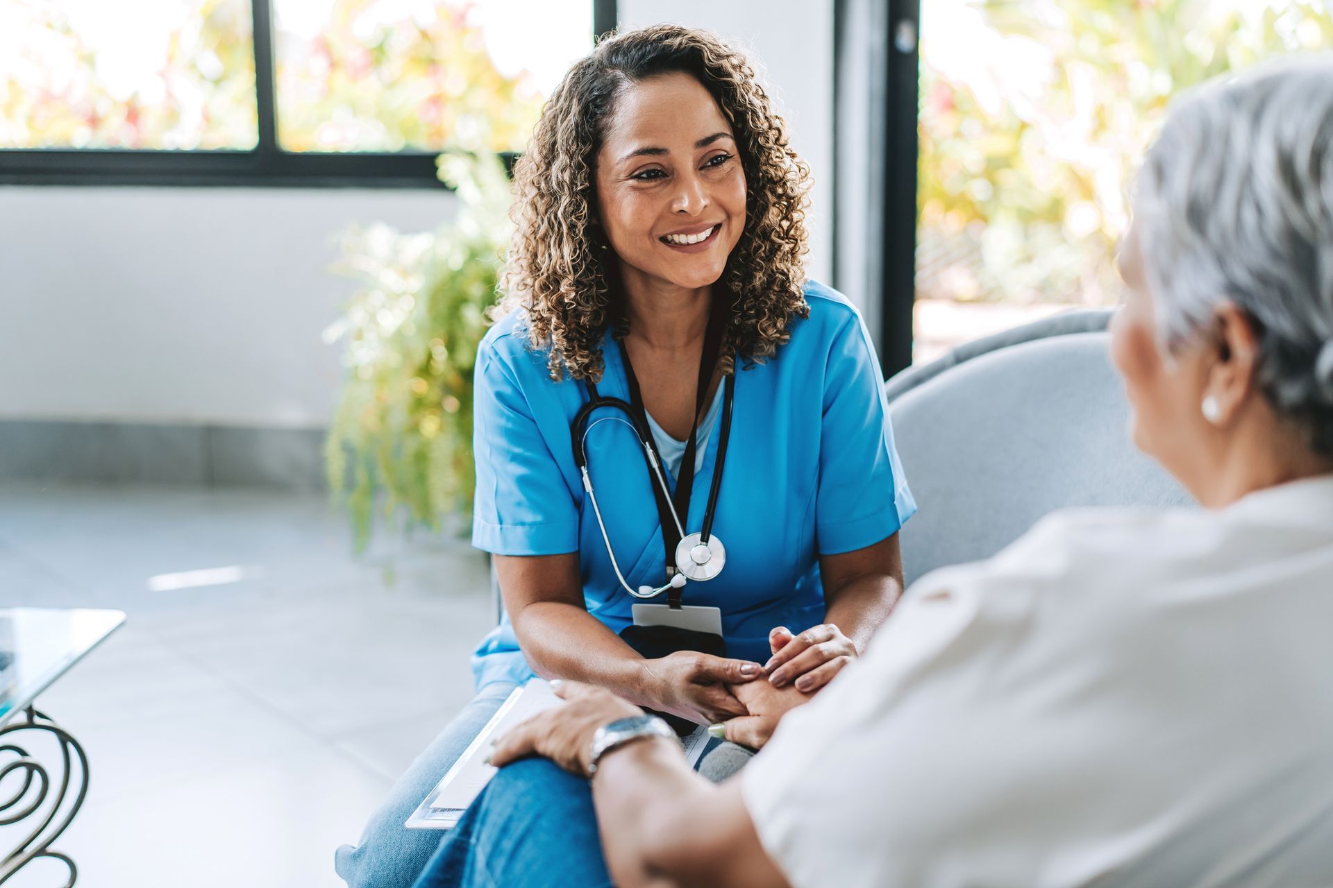 Nurse in blue scrubs smiling while holding a patient’s hand; interior setting.
