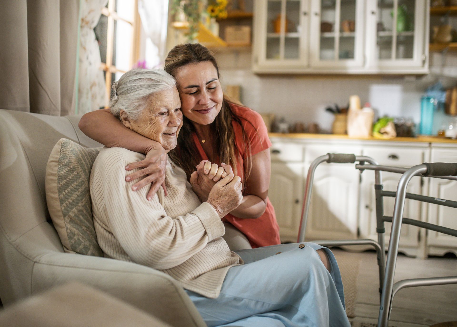 Woman hugging and holding hands with older woman in a home setting; walker nearby.