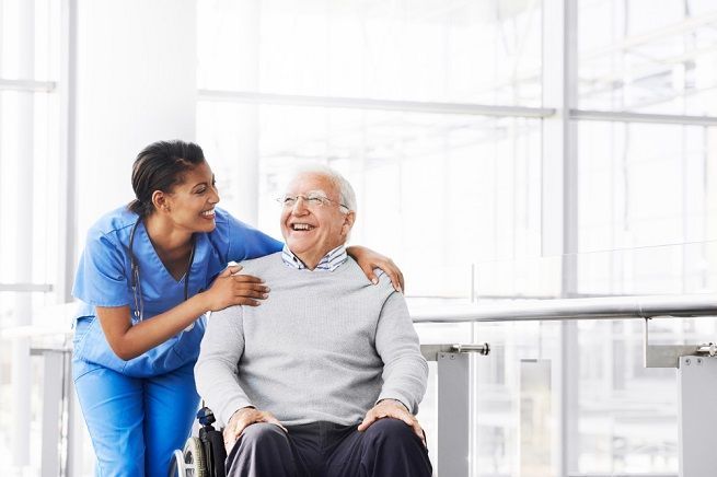 Nurse in blue scrubs with arm around smiling person in a wheelchair indoors.