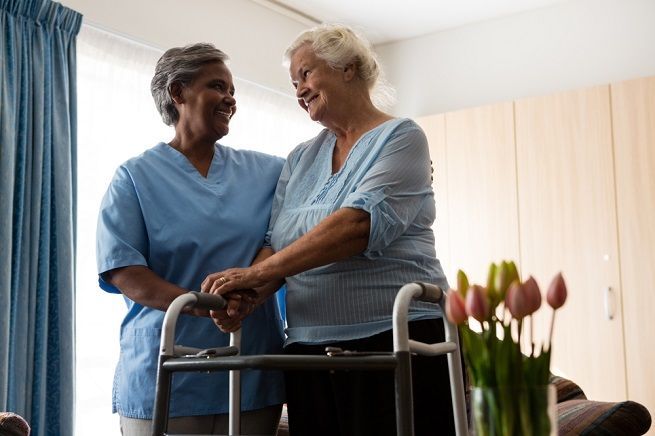 Caregiver assisting an older person using a walker. Both smile warmly. Indoors with flowers.