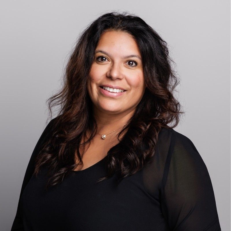 Een vrouw met lang donker haar lacht, gekleed in een zwart topje tegen een grijze achtergrond.