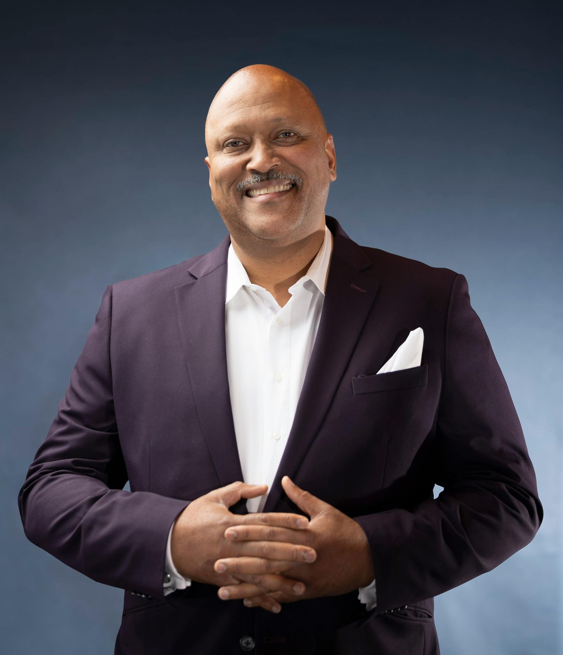 Man in paars pak lacht en houdt zijn jasje open; een wit pochet is zichtbaar; blauwe achtergrond.