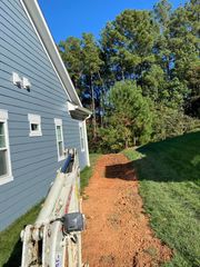 A construction vehicle sits on a path of exposed red soil beside the blue siding of a house, bordering a wooded area.