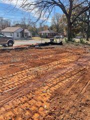 A view of a plowed, muddy field with tire tracks in the foreground, facing houses and trees under a sunny sky.