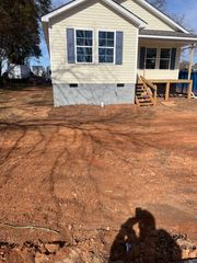 A beige house with blue shutters sits on a grey concrete foundation with an unfinished front porch and bare dirt yard.