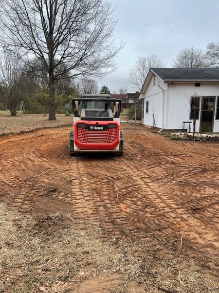 A red and white Bobcat skid steer loader sits on a dirt lot in front of a white house on a cloudy day.