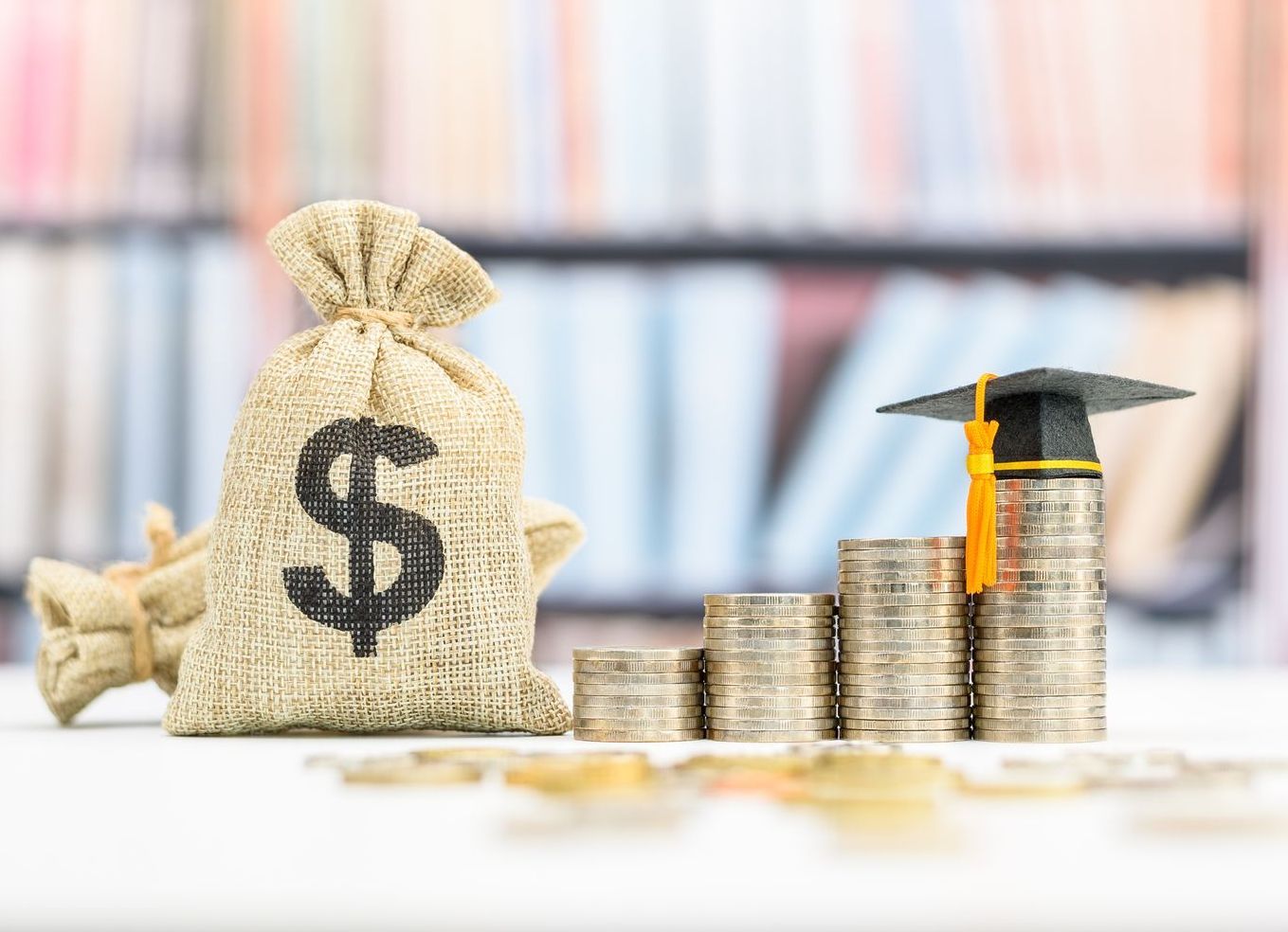 A stack of coins next to a bag of money and a graduation cap.