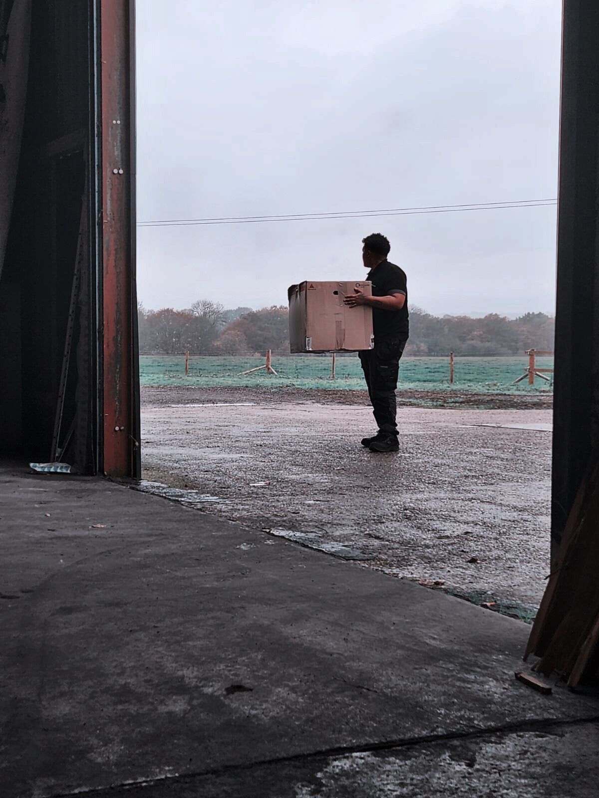 Person carrying a cardboard box outside a large open doorway in the rain.