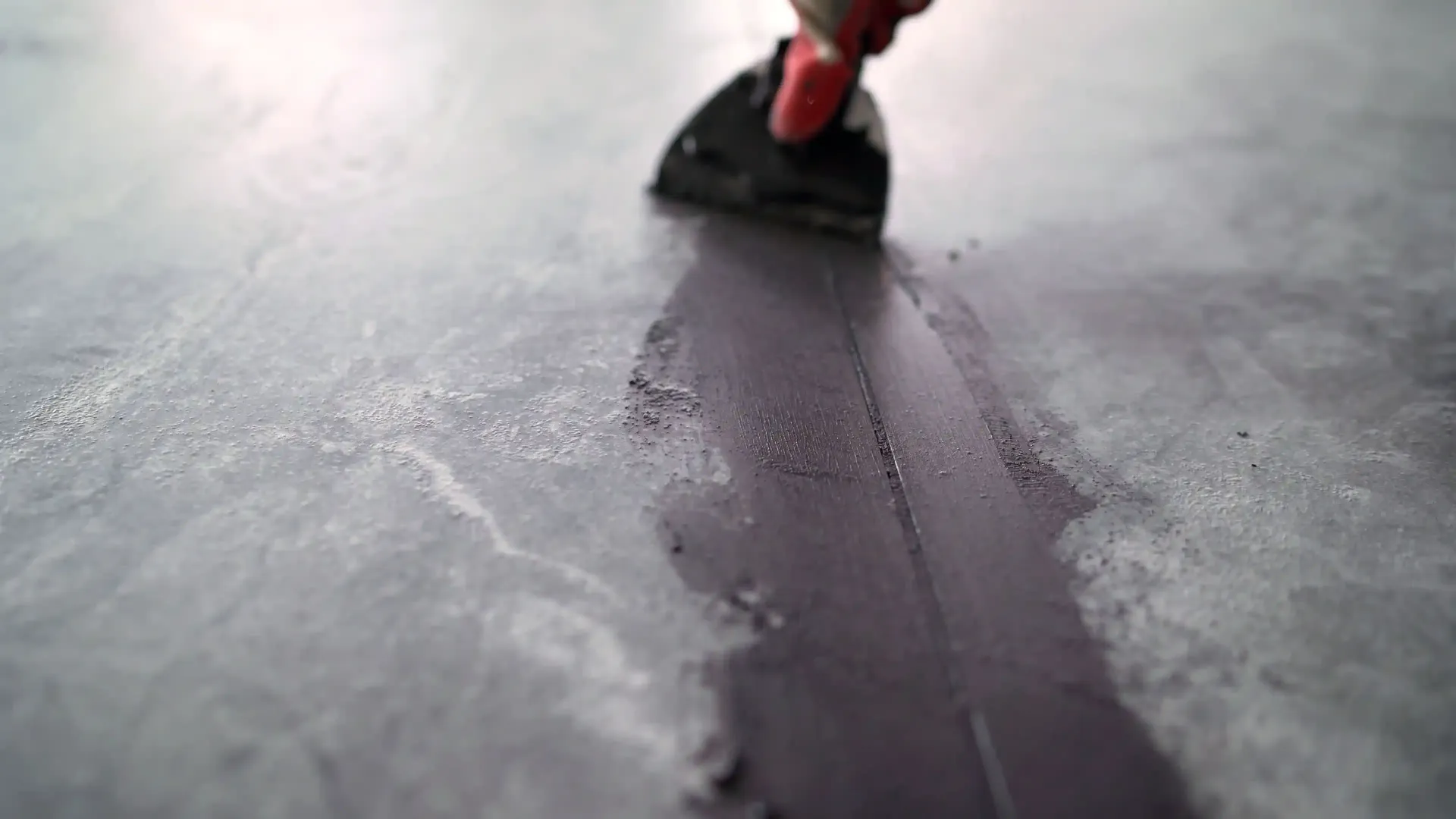 Person using a trowel to smooth dark grey material on a concrete floor.