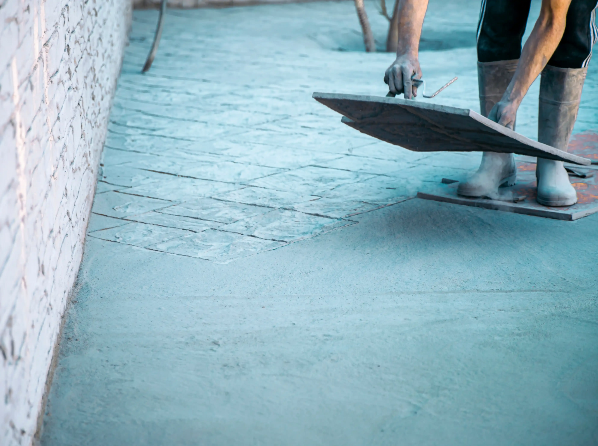 Person smoothing concrete with a trowel, wearing boots, next to a white wall.